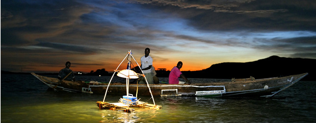 Kenianische Fischer auf einem Boot im Dunkeln, auf einer Schwimminsel davor eine elektrische Lampe.